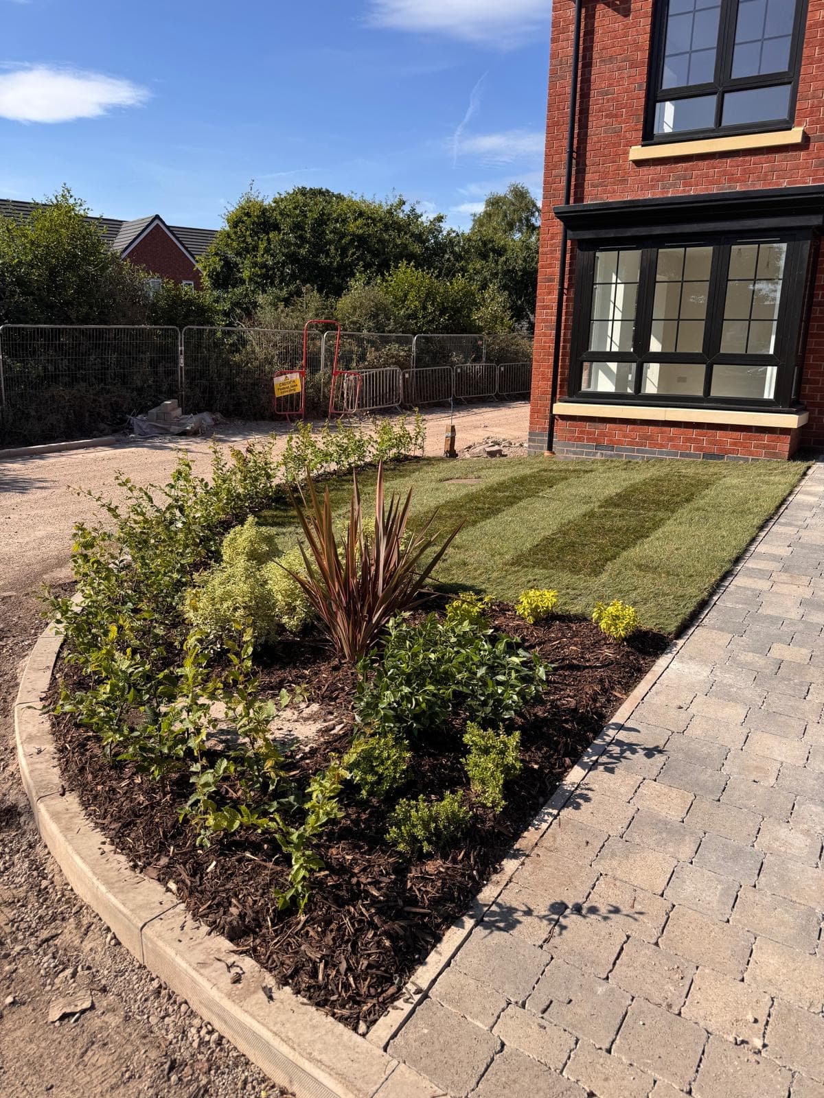 A rounded bed of young shrubs and a red-leaved phormium in bark mulch beside new turf and block paving, in front of a red-brick house with black-framed windows