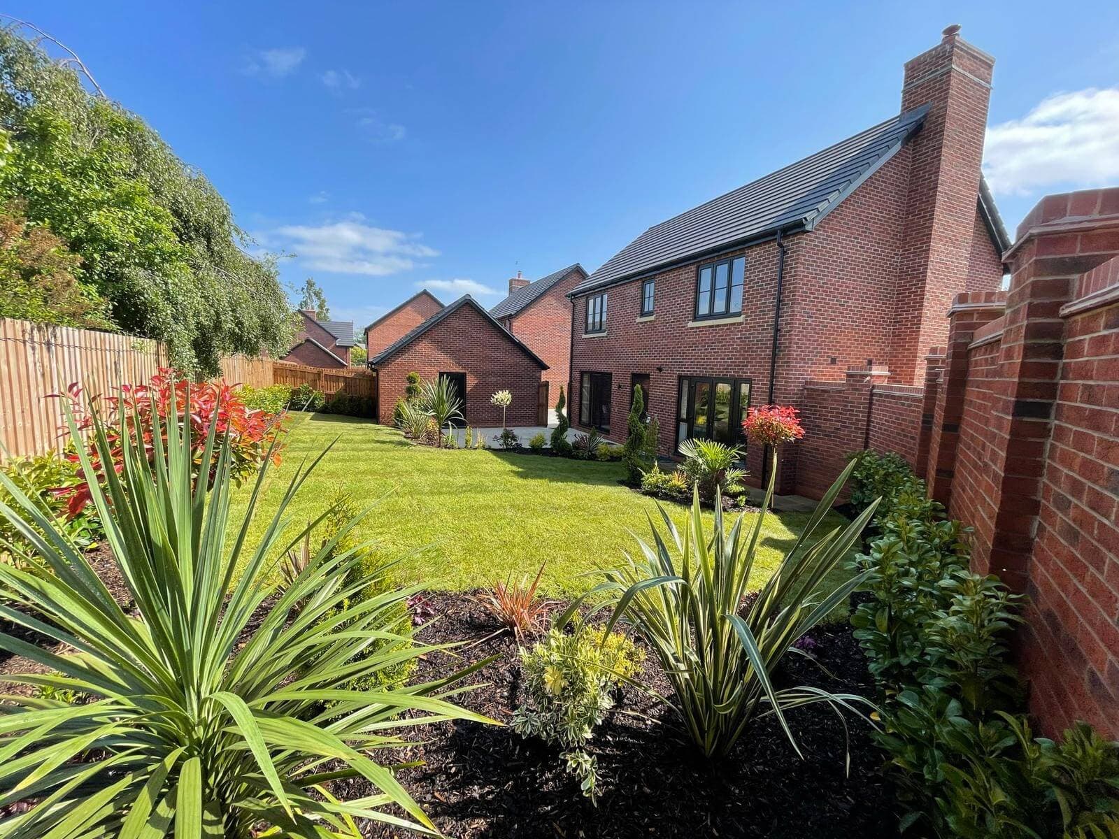 A new-build back garden planted and turfed: spiky yuccas and red-leaved shrubs in dark mulch, a striped lawn and the brick house behind