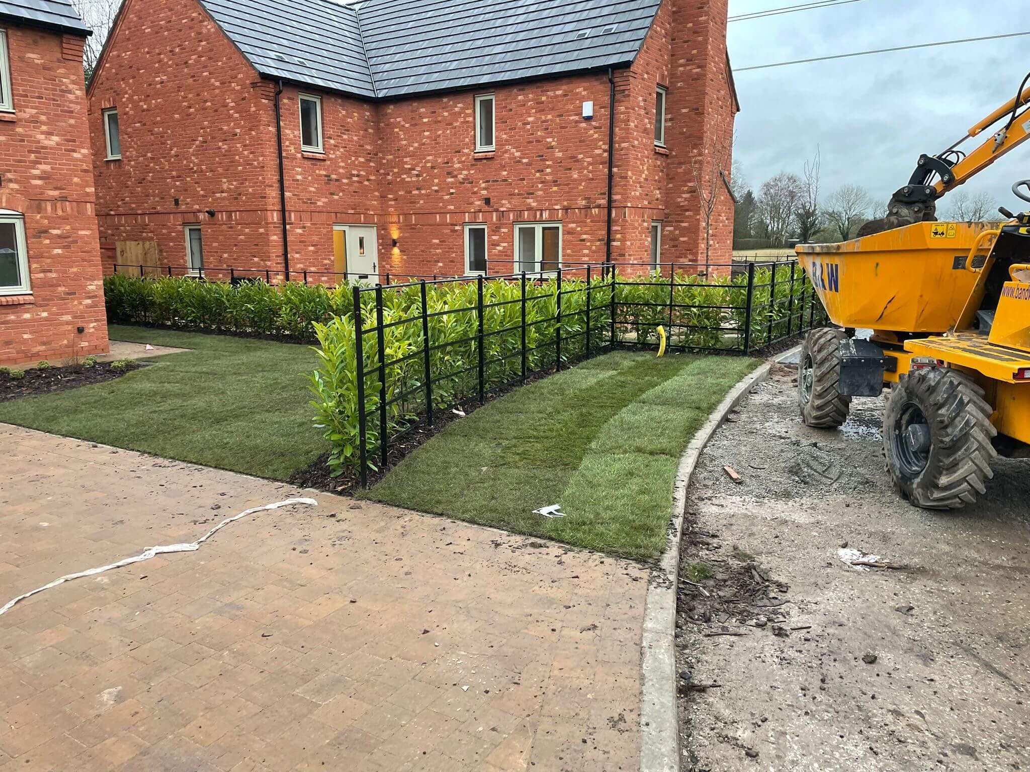Newly laid turf and a young evergreen hedge behind black iron railings in front of a red-brick new-build house, with a yellow dumper truck on the unfinished road beside them