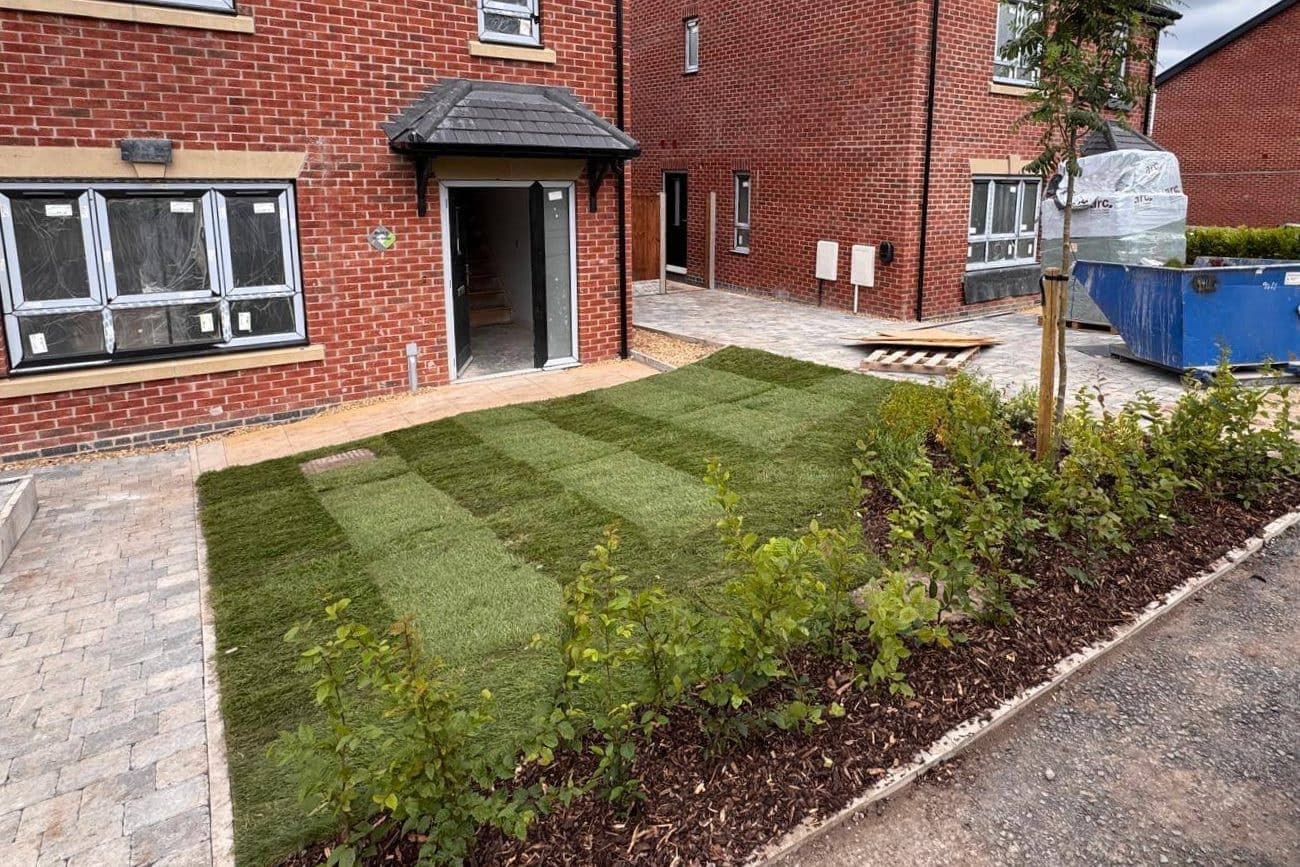 A new front garden of striped turf and a bark-mulched bed of young hedgerow shrubs in front of red-brick houses, with a skip and pallets on the development behind