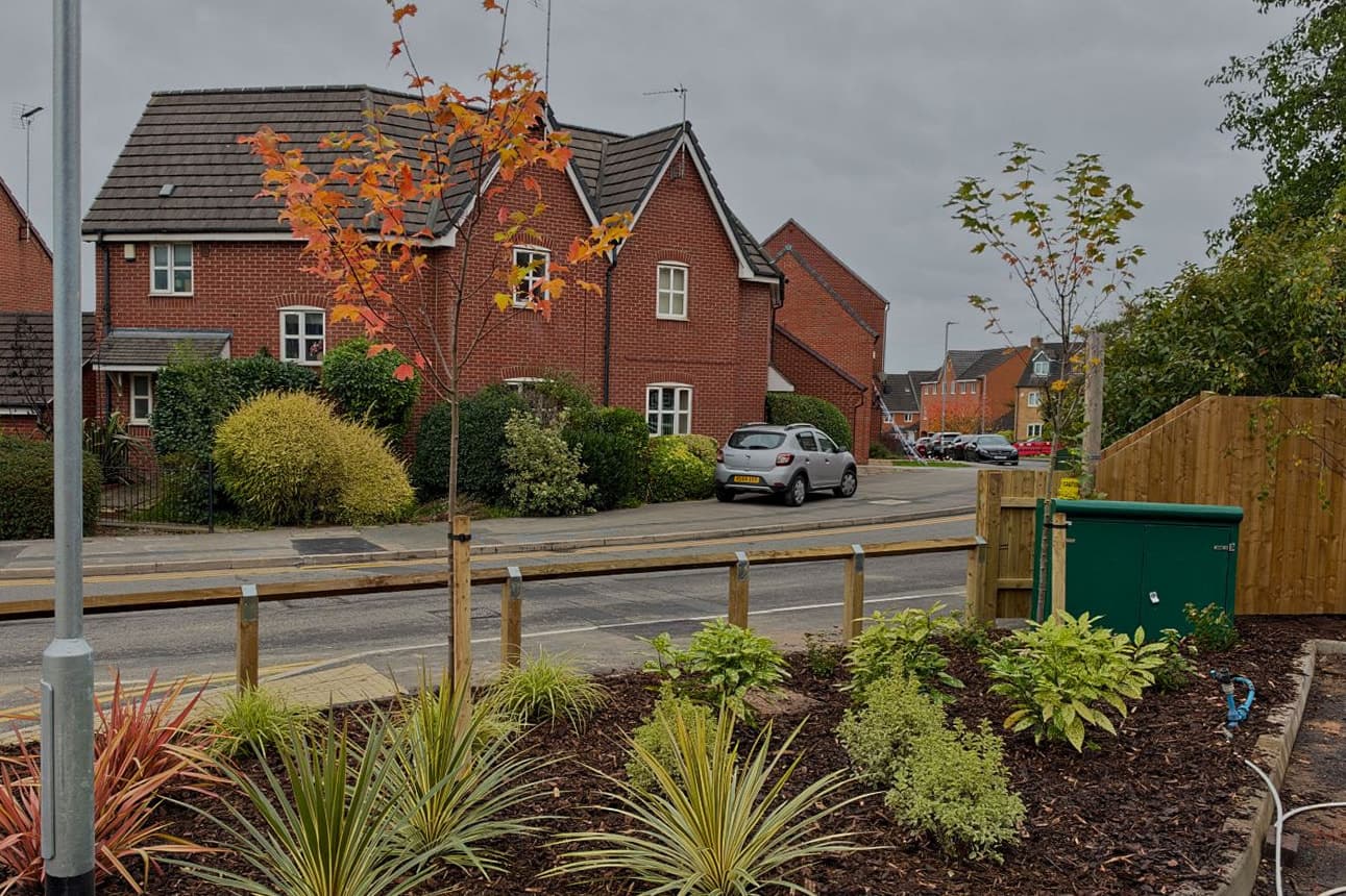 A new roadside bed in bark mulch with red and green phormiums, striped cordylines and golden shrubs, a young maple in autumn colour on a stake and a timber knee rail, with red-brick houses across the road